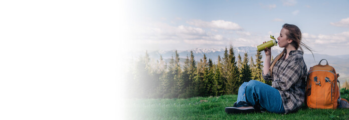 A young girl with loose hair in a shirt and jeans with an orange backpack drinks tea from a thermal mug sitting on the green grass against the backdrop of the mountains. photo on horizontal banner