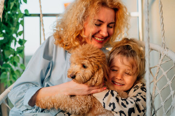 close-up portrait happy mother with daughter at home on the balcony, sitting on a hammock chair, cuddling with a pet: a poodle dog on a warm sunny evening at sunset during the golden hour