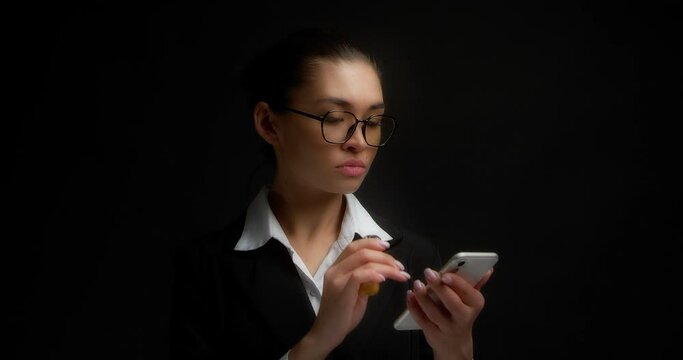 Brunette with gathered hair, glasses and office clothes is standing on an isolated black background, flipping through a news feed in a messenger and smoking, vaping an electronic cigarette.