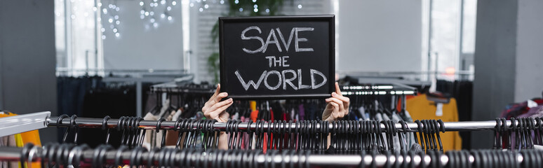 Cropped view of saleswoman holding board with save the world lettering near hangers in vintage...