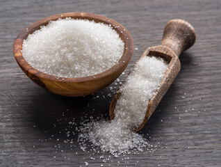 White refined sugar in wooden bowl and spoon on dark wooden table.