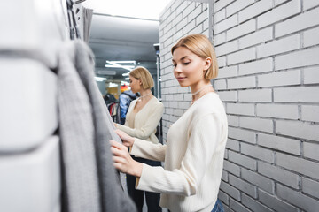 Young customer looking at blurred clothes in dressing room in second hand.
