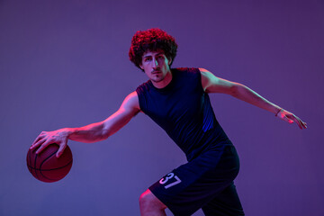 Studio shot of young muscular basketball street player dribbling ball isolated on purple background...
