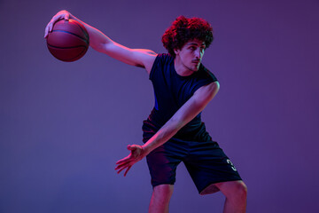 Dynamic portrait of professional basketball player jumping with ball isolated on purple background...