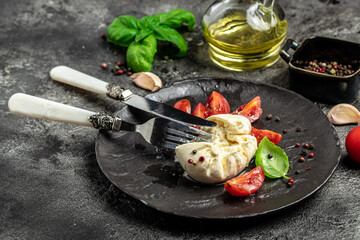 Salad with creamy Italian Burrata Cheese Served with Olive Oil, Basil Leaves and cherry tomatoes on white plate on a dark background. Restaurant menu, dieting, cookbook recipe top view