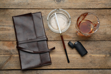 Glass ashtray with long cigarettes holder, lighter, clutch and alcohol drink on wooden table, flat lay