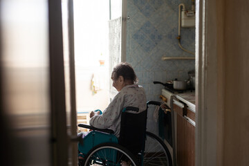Old lonely woman sitting in a wheelchair near the window in his house - obrazy, fototapety, plakaty