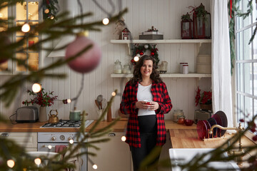 a young beautiful pregnant woman stands in the kitchen in cozy clothes and holds a mug in her hands. next to it is a decorated Christmas tree with lights. concept of future motherhood