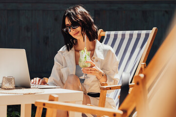 Young Brunette Woman in Sunglasses Working on Laptop and Drinking Cocktail While Sitting on the Terrace of the Restaurant, Freelancer at Work