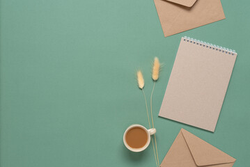 Home office desk. Notepad, cup of coffee, brown envelopes and dry lagurus grass. Green pastel paper background. Top view, flat lay.