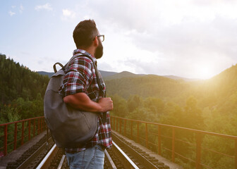 A young tourist with a beard and glasses in a plaid shirt with a backpack stands on the railway...