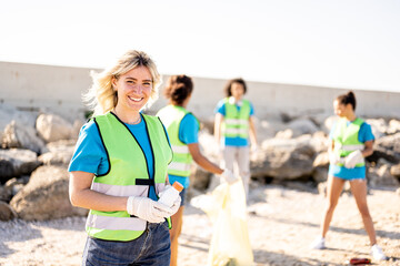 Young blonde woman smiling at the camera while her teamwork cleaning the rubish on a beach,...