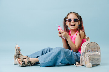 Cheerful tween girl in pink shirt and round sunglasses using her phone. Sitting on the floor over...
