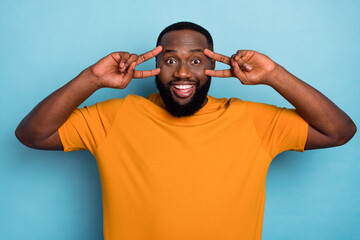 Photo of cheerful pretty guy wear orange t-shirt v-signs cover eyes isolated blue color background