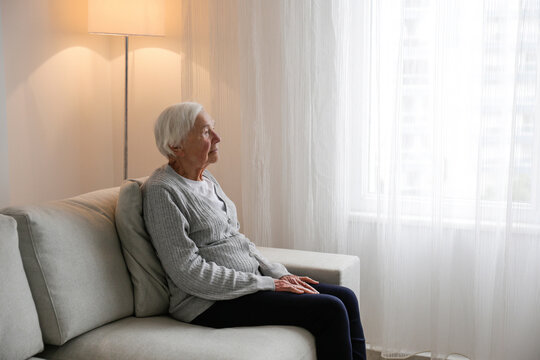Portrait of elderly lady alone at home. Senior woman 86 years of age sitting on the couch by the window in her apartment. Background, copy space, close up.