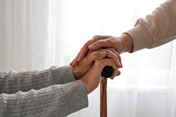 Senior woman holding quad cane handle in elderly care facility. Female personnel comforting mature lady in nursing home, sitting with walking stick. Background, close up, cropped shot.