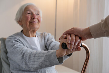 Senior woman holding quad cane handle in elderly care facility. Unrecognizable female comforting mature lady in nursing home, sitting with walking stick. Background, close up, cropped shot. - obrazy, fototapety, plakaty