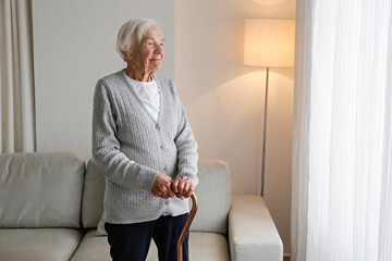 Senior woman 86 years of age standing by the window with walking cane in her apartment. Portrait of...