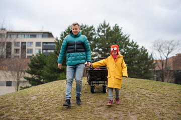 Father with his little daughter with Down syndrome pulling trolley outdoors in park.