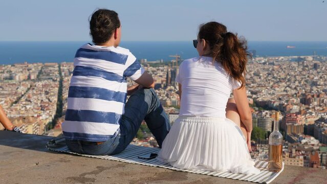 Man and woman drink wine at height of viewpoint, make sip from glass, view from back. Scenic aerial panorama of Barcelona seen down around. Two people rest at summer evening