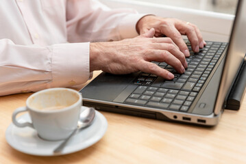 close-up of an unrecognizable older man's hands typing on the keyboard of a laptop, a cup of coffee by his side