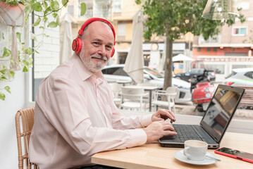 smiling older man sitting in a coffee shop with headphones uses laptop to learn languages, online classes, enjoying his retirement, looking at the camera