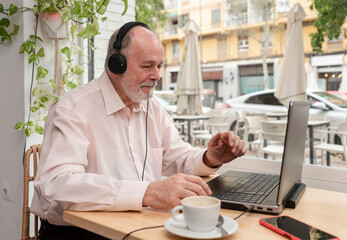 a retired man sitting in a coffee shop with headphones using his laptop to take online classes for seniors while drinking his coffee