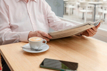 an unrecognizable man reading a daily newspaper, his cup of coffee next to him and a cell phone