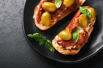 Bruschetta with sun dried tomatoes. Toast or sandwich with ricotta, sun dried tomatoes and Provence herbs with basil servered on black plate on black background table. Selective focus.