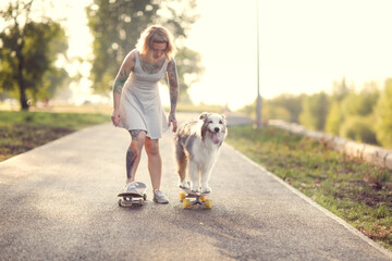 Naklejka na ściany i meble Cute red-haired hipster girl with a tattoo rides a skateboard with an Australian Shepherd dog on the sidewalk in the park, warm summer evening