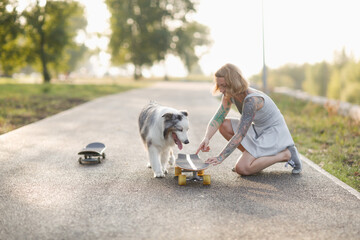 Naklejka na ściany i meble Cute red-haired hipster girl with a tattoo rides a skateboard with an Australian Shepherd dog on the sidewalk in the park, warm summer evening