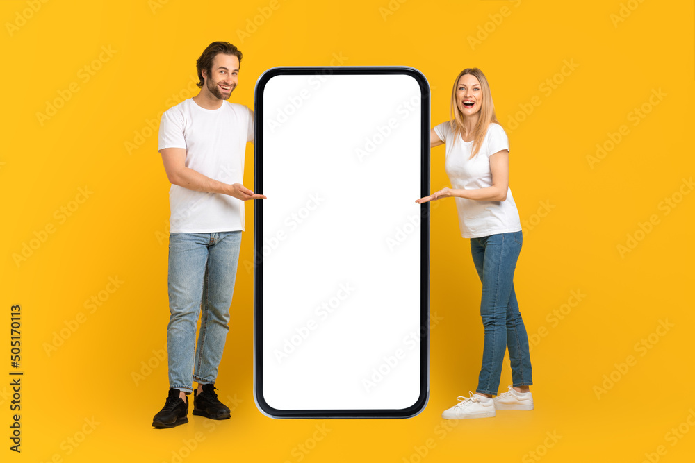 Wall mural Happy young caucasian male and female in white t-shirts presentation huge phone with empty screen