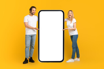 Happy young caucasian male and female in white t-shirts presentation huge phone with empty screen