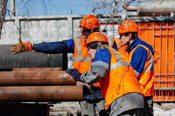 Team of construction workers in helmets discusses place of laying pipes. Three workers confer on...