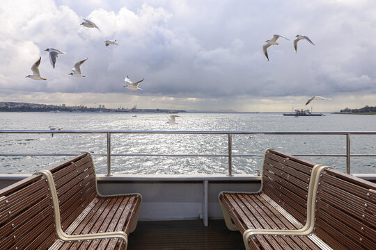 Ferry Across The Bosporus In Istanbul, Turkey. Seagulls Soar In The Cloudy Spring Sky. Two Rows Of Empty Benches Facing Each Other.