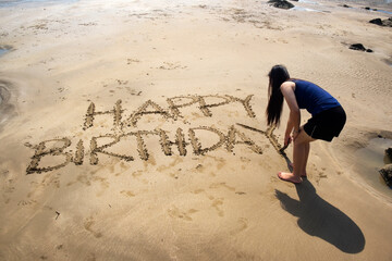 Rear view of woman writing happy birthday on beach sand with a wooden stick.