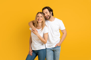 Fototapeta premium Glad young european guy and woman in white t-shirts hugging, have fun, isolated on yellow background