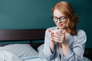 young red-haired woman in glasses and pajamas sits on the bed in the morning and drinks coffee