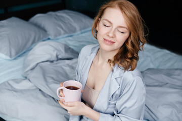 young red-haired woman and pajamas sits on the bed in the morning and drinks coffee