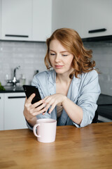 young caucasian red-haired woman in pajamas in the morning in the kitchen drinks coffee and checks messages on her smartphone