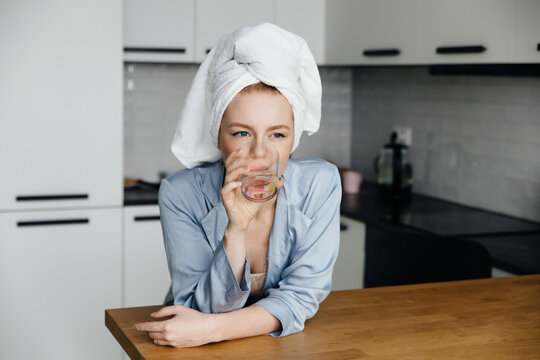 Young woman dressed in pajamas drinking water with a piece of lemon and standing in her kitchen.