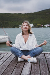 Fototapeta na wymiar Adorable young woman sits on pier on beautiful mountain lake and meditates