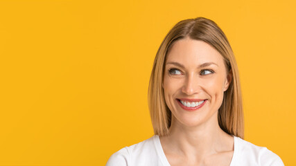 Smiling cheerful young european lady blonde in white t-shirt looks at free space, isolated on yellow background
