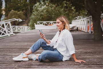 Charming young woman sits on pier on beautiful mountain lake and communicates on mobile phone