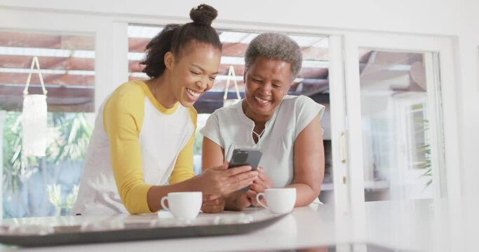 African american mother and daughter smiling while using smartphone together at home