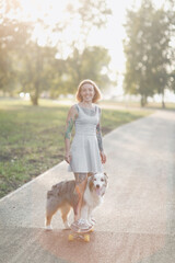 Cute hipster woman girl with tattoo rides skateboard with an Australian Shepherd dog on sidewalk in park, warm summer evening, training dog