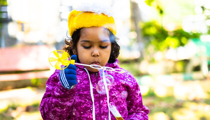 Close up shot of serious girl kid with winter wear on swing blowing soap bubble at park - concept...