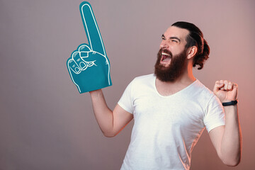 Excited young bearded man wearing blue foam fan glove is shouting loud. Studio shot over grey...