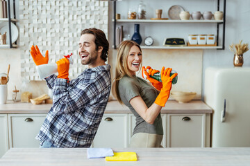 Satisfied man with stubble in rubber gloves with spray and wife sing at imaginary microphone in...