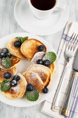 On a white plate are pancakes with blueberries and mint, cutlery and a cup of tea on a light table. Tasty food.